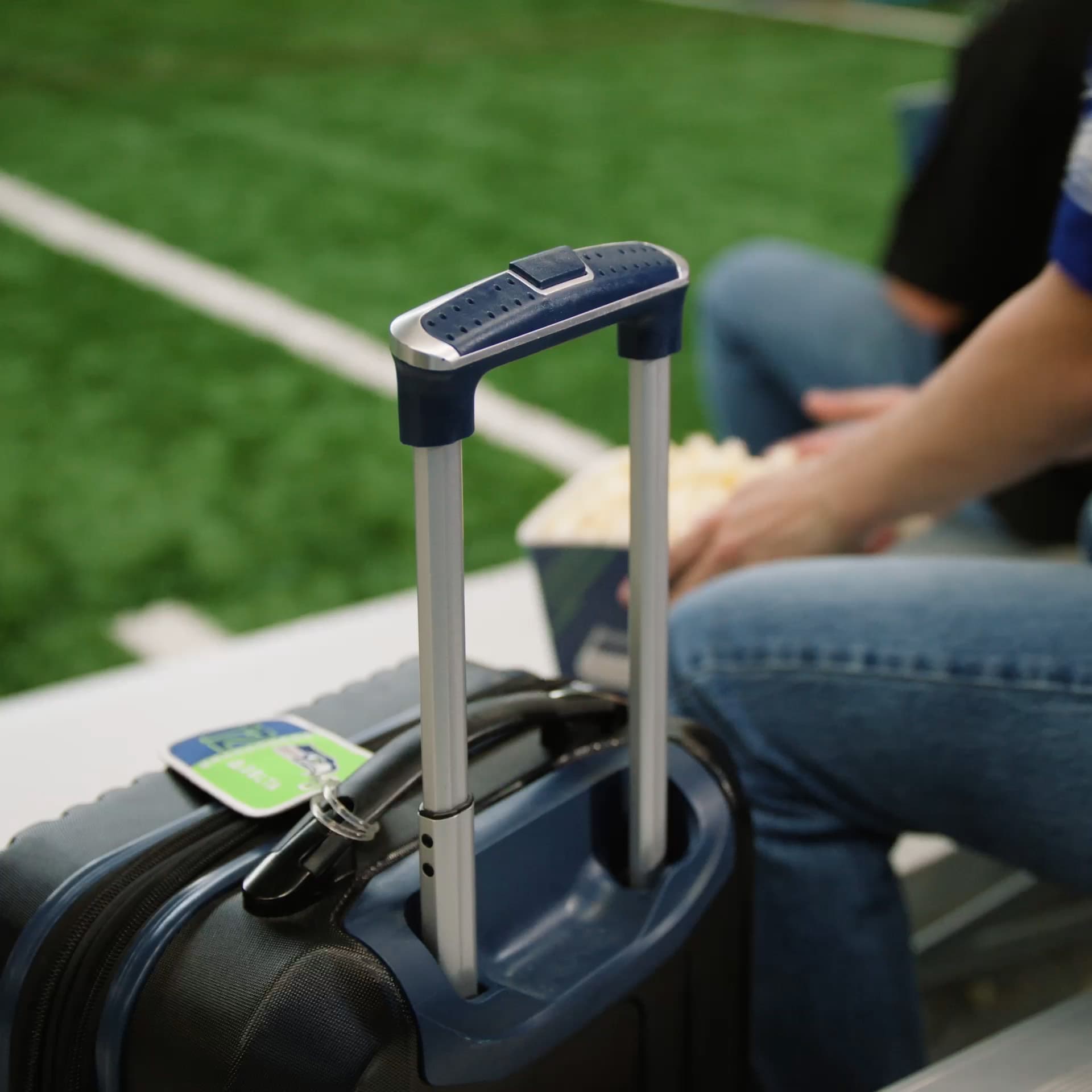A Seahawks-branded luggage tag on a rolling suitcase, with a fan seated behind it beside the field