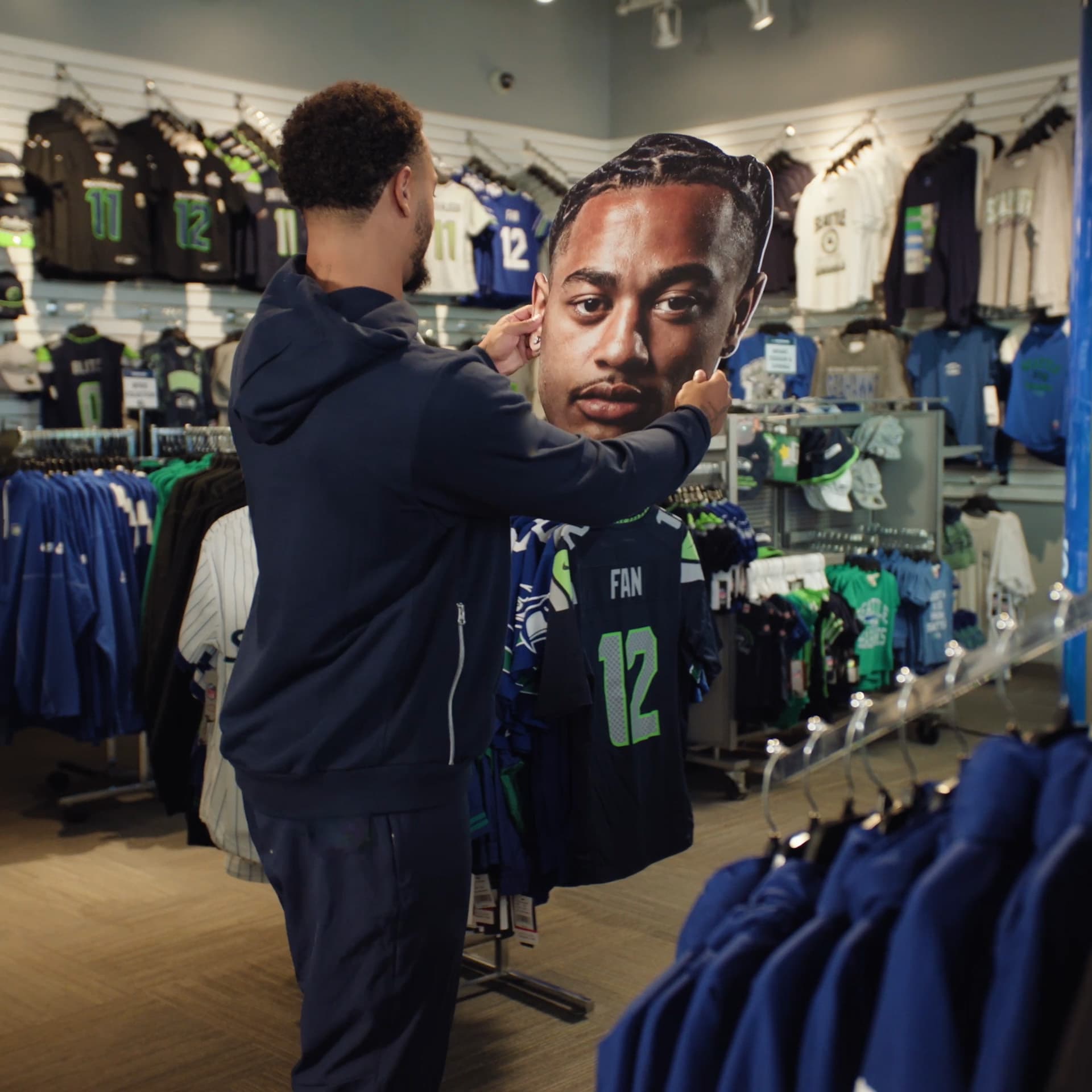 A fan in Seahawks gear browsing jerseys in the team Pro Shop beside a life-size player cutout