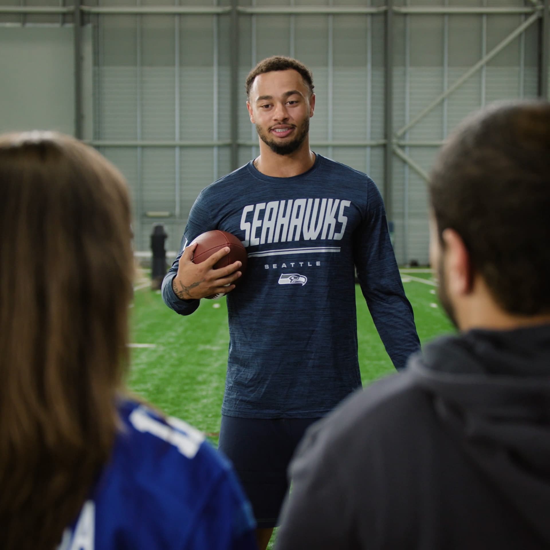 A Seahawks player in a team practice shirt holding a football, talking with two fans on an indoor practice field